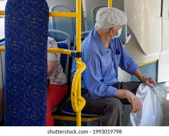 An elderly man in a blue shirt and beige cap sits in the passenger seat facing forward in the bus. A medical mask is visible on his face, and he holds a white bag in his left hand.