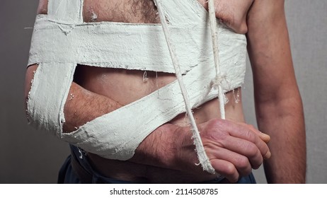 Elderly Man With Broken Forearm Fixed With Plaster Cast And White Bandage On Bare Torso Stands Straight By Grey Wall At Home Closeup.