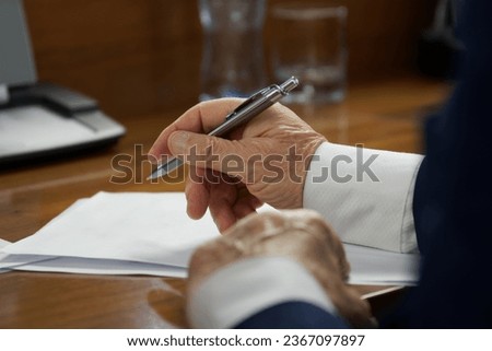 Elderly man in business suit, sitting at a table with pen, fills out an agreement or will form. Concept of writing a will, concluding contract or filling out forms. Photo. Selective focus. Close-up