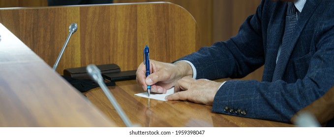 An Elderly Man - A Businessman, Lawyer Or Teacher Writes Something On A Piece Of Paper, Sitting At A Table Or Desk. Meeting, Negotiation Or Court Hearing. No Face