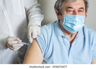 An Elderly Man Of Caucasian Appearance In A Mask Receives A Dose Of Coronavirus Vaccination At Home, The Doctor Came To The Patient To Give A Protective Injection Against The Disease