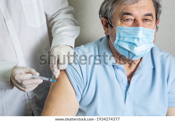 An\
elderly man of Caucasian appearance in a mask receives a dose of\
coronavirus vaccination at home, the doctor came to the patient to\
give a protective injection against the\
disease
