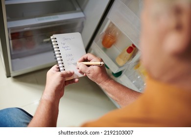 Elderly Man Checking His Empty Fridge And Writing Down Shopping List In Notepad