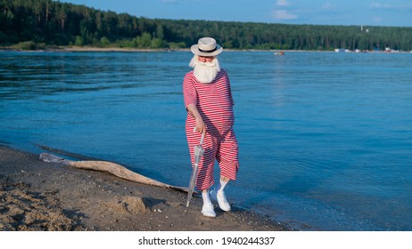 An Elderly Man In A Classic Bathing Suit Walks Along The Beach With An Umbrella On A Hot Summer Day