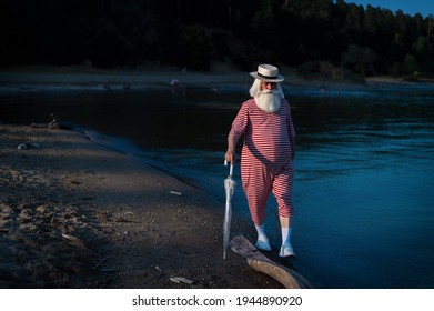 An Elderly Man In A Classic Bathing Suit Walks Along The Beach With An Umbrella On A Hot Summer Day