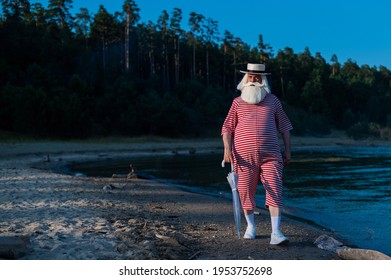 An Elderly Man In A Classic Bathing Suit Walks Along The Beach With An Umbrella On A Hot Summer Day