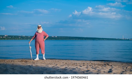 An Elderly Man In A Classic Bathing Suit Walks Along The Beach With An Umbrella On A Hot Summer Day