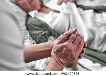 elderly man crying and mourning the loss of his wife, sitting by her side. focus on hands. coronavirus, covid-19 concept