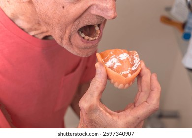An elderly man with decayed teeth prepares to insert his upper denture, which is coated with adhesive powder. Tooth loss in advanced age, dental health, and prosthetic solutions.