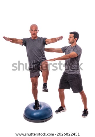 An elderly man doing a balance exercise, on one leg, on a hemisphere ball. With help of a fitness trainer. On a white isolated background.