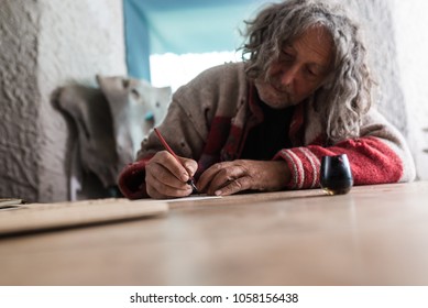 Elderly man doing calligraphy using a nib pen and ink or pigment from a pot in a low angle tilted view across a wooden table with foreground copy space.
