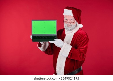 Elderly man dressed as Santa Claus pointing at a blank laptop screen with a red backdrop. Green screen ready for customization allowing for various design options - Powered by Shutterstock