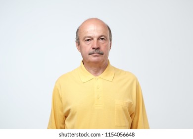 Elderly Man Emotions, Portrait Of Serious Senior Caucasian Man In Yellow T-shirt Looking At Camera Against Gray Wall