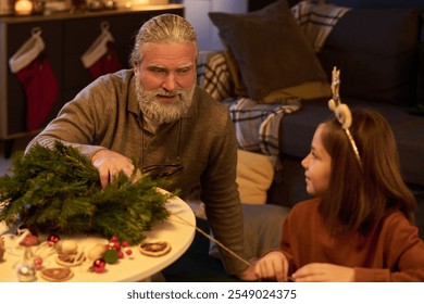 Elderly man engaging with young girl while making holiday decorations, creating festive atmosphere in living room. Christmas stockings and decorations visible in background - Powered by Shutterstock