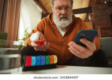 Elderly man examining pill bottle while looking at smartphone. Using pill organizer. Online med services. Concept of health and medical care, aging, medicine, treatment - Powered by Shutterstock