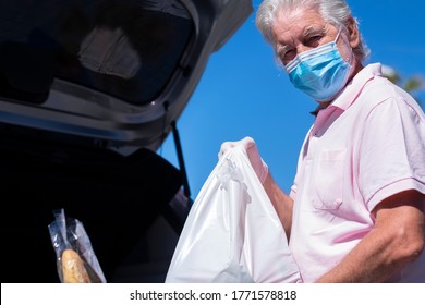 Elderly Man With Face Mask Loads Shopping Bags Into The Trunk Of The Car, Outdoor Parking In The Sun - Concept Of Active Retired Senior People