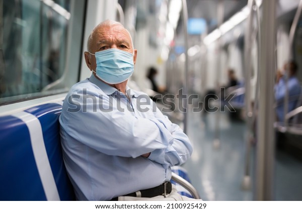 Elderly man in face mask sitting on bench\
inside subway car and waiting for his\
stop.