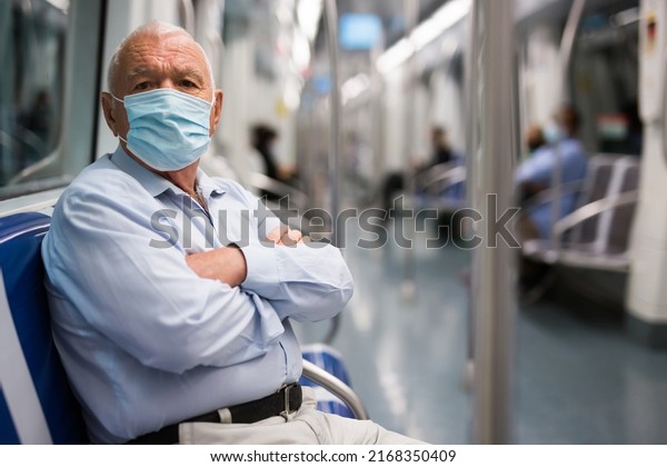 Elderly man in face mask sitting on bench\
inside subway car and waiting for his\
stop.