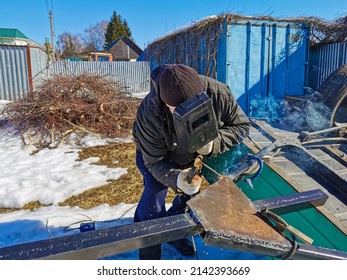 An elderly man foreman repairs a car trailer using electric arc welding. A worker sparks while welding steel on a spring sunny day against the backdrop of snow.