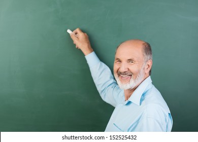 Elderly Man With A Friendly Smile Writing On A Blank Chalkboard During A Business Presentation Or While Teaching Class At College