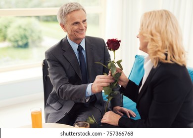 An elderly man gives a rose to an elderly woman. They are sitting on the couch. On the table are glasses with orange juice.