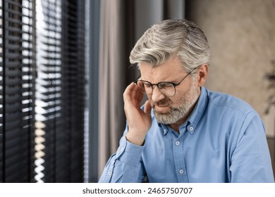 Elderly man with glasses and grey hair deep in thought by a window with blinds, in a calm contemplative mood - Powered by Shutterstock