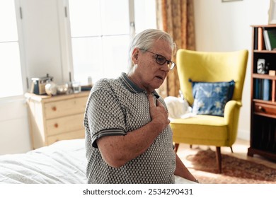 Elderly man with glasses holding chest, sitting in a cozy room. Concerned expression, wearing a patterned shirt. Bright, comfortable setting with a yellow chair. Man with chest pain, heart attack. - Powered by Shutterstock