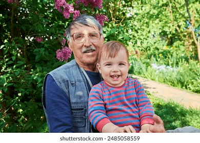 An elderly man with glasses and a mustache holds a happy, smiling toddler on his lap. They are sitting in a garden with vibrant green foliage and blooming purple flowers in the background. - Powered by Shutterstock