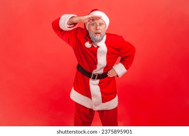 Elderly man with gray beard in santa claus costume keeping palm over head and looking attentively far away, peering with expectation at long distance. Indoor studio shot isolated on red background. - Powered by Shutterstock