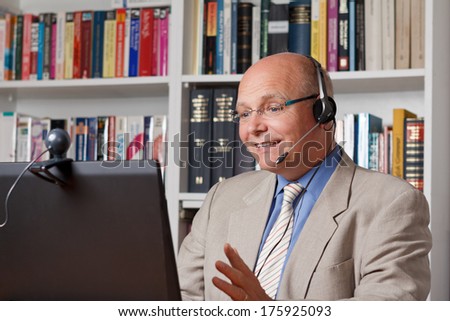 Elderly man with headset, computer and webcam smiling while having an online video chat, live customer support service, telemedicine or telelearning concept