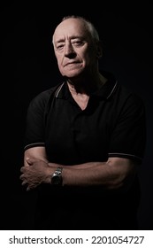 An Elderly Man In His 70s Stands With Folded Arms And Looks Calmly At The Camera. Studio Portrait On A Black Background.