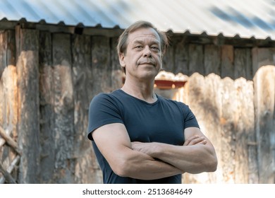 An elderly man with his arms crossed poses in front of a rustic wooden building.  - Powered by Shutterstock