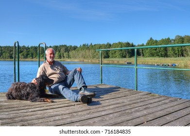 An Elderly Man And His Dog Are Sitting On The Pier And Enjoying The Beautiful Day At The Lake.