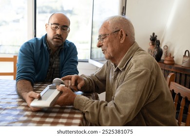 an elderly man in his eighties with his son-in-law changing the batteries of a meteorological device and watching how it works sitting at a table - Powered by Shutterstock