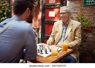 An elderly man and his young friend enjoying a game of chess in a cheerful atmosphere in the bar. Leisure, bar, friendship, outdoor - Powered by Shutterstock