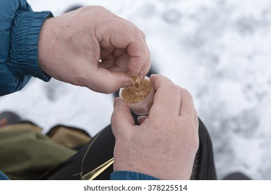 An Elderly Man Is Ice Fishing And He Is Picking A Bait From The Plastic Cup.