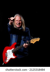 elderly man with long gray hair in blue denim shirt plays the electric guitar on black background