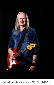 elderly man with long gray hair in blue denim shirt plays the electric guitar on black background