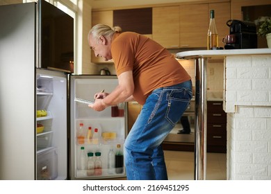 Elderly Man Looking At Opened Fridge And Writing Down What To Buy In Grocery Store