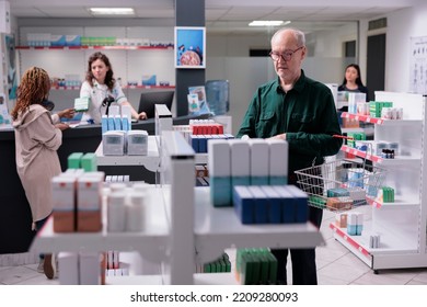 Elderly Man Looking At Shelves Full With Pharmaceutical Products, Buying Supplements After Reading Medication Leaflet. Clients Holding Baskets Putting Pills Package In It. Health Care Service