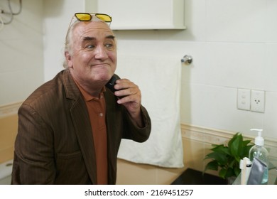 Elderly Man Making Funny Faces When Shaving Off Beard In Front Of Bathroom Mirror