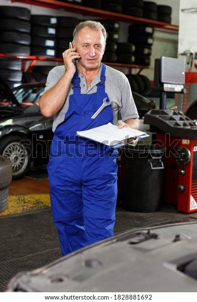 Elderly man mechanic discussing with\
client on phone upcoming repair of car in auto\
workshop