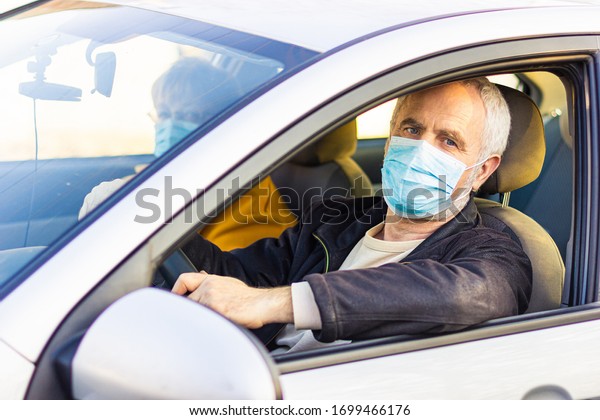 An elderly man in a medical\
face mask driving a car. Coronavirus pandemic concept. Road trip,\
travel and old people concept - happy senior couple driving in\
car