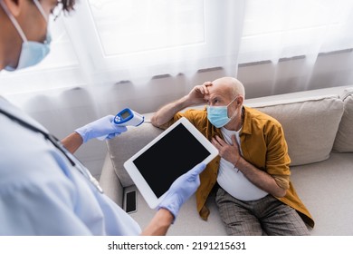 Elderly Man In Medical Mask Sitting Near Blurred African American Nurse With Pyrometer And Digital Tablet At Home