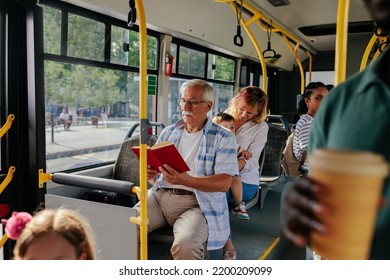 An Elderly Man Is On The City Bus Reading A Book