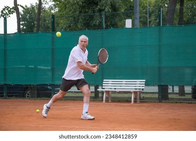 An elderly man playing tennis is hitting a backhand. The moving at camera. Open ground. - Powered by Shutterstock