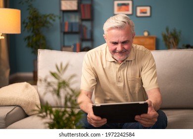 An Elderly Man Plays On A Tablet While Sitting On The Living Room Couch At Home In The Evening. Older Man Playing Games, Cards On Tablet. Man Sitting On Couch In Living Room Happy With Online Shopping