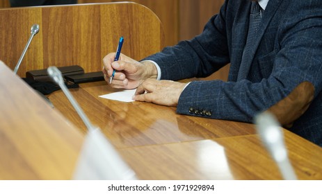 An Elderly Man - Politician, Lawyer Or Teacher Writes On A Piece Of Paper While Sitting At A Table In The Boardroom. A Court Hearing Or Meeting Of Politicians And Officials. No Face