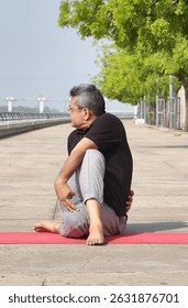 Elderly man practicing Ardha Matsyendrasana - Half Lord of the Fishes Pose on a yoga mat in a outdoor setting, promoting spinal flexibility, detoxification, and inner calm at riverfront ahmedabad.