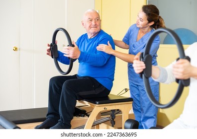 Elderly man practicing pilates with magic circle in rehabilitation center to improve and maintain mobility under supervision of qualified female doctor. Therapeutic physical training